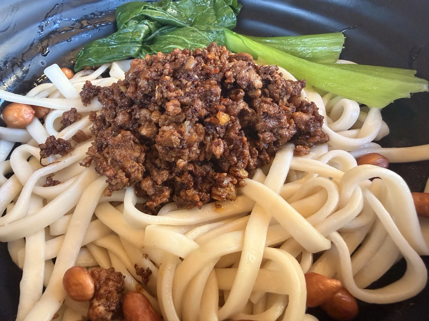 Wide noodles with a savoury topping, peanuts, and leafy greens
