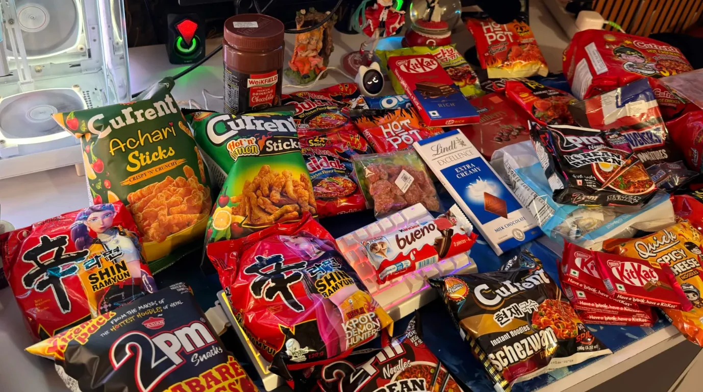 Packets of noodles, crisps, and chocolate spread across a computer desk