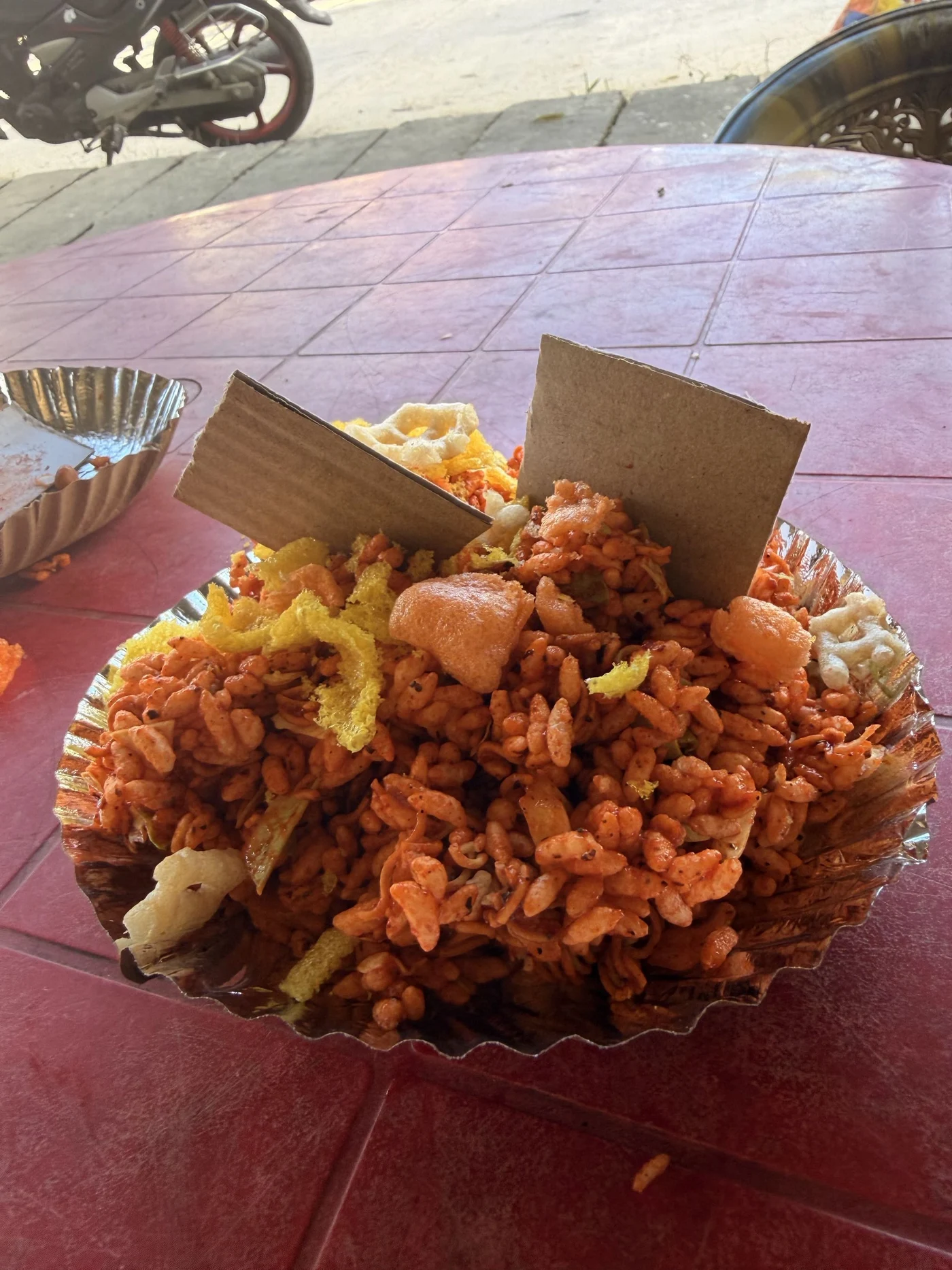 A foil plate piled with a spicy puffed-rice snack on a pink table